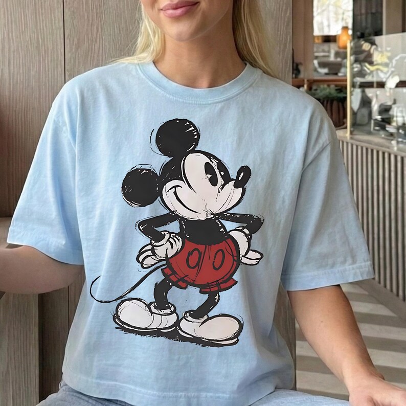 May include: Light blue t-shirt featuring a distressed graphic of Mickey Mouse. The cartoon character is depicted in black, white, and red, with a classic pose. The shirt has short sleeves and a crew neck.