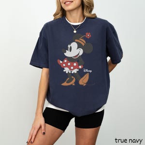 Może przedstawiać: Granatowa koszulka z grafiką Myszki Minnie. Postać z kresk&oacute;wki ma na sobie czerwoną sukienkę w kropki i kapelusz z kwiatkiem. Logo Disneya jest wydrukowane poniżej. Koszulka ma luźny kr&oacute;j.