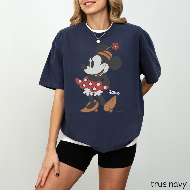 Może przedstawiać: Granatowa koszulka z grafiką Myszki Minnie. Postać z kresk&oacute;wki ma na sobie czerwoną sukienkę w kropki i kapelusz z kwiatkiem. Logo Disneya jest wydrukowane poniżej. Koszulka ma luźny kr&oacute;j.