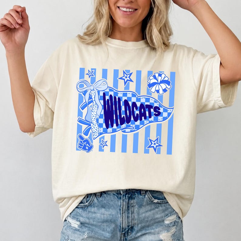 May include: A cream-colored short-sleeve t-shirt featuring a blue graphic print. The design includes a checkered pennant with the text "WILDCATS" in dark blue, surrounded by blue striped patterns, stars, a cheerleading pom-pom, and a bow.