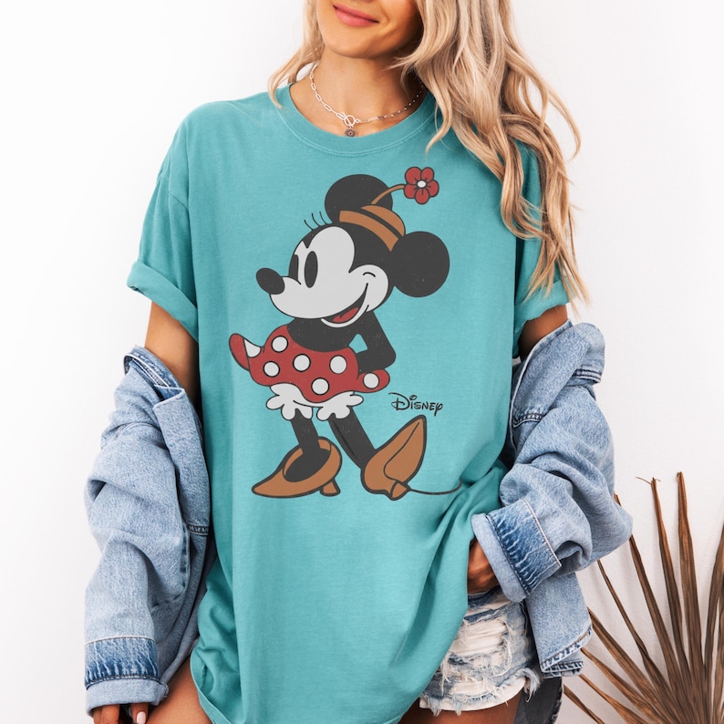 Może przedstawiać: Turkusowa koszulka z Myszką Minnie. Grafika przedstawia Minnie w czerwonej sukience w kropki, brązowych butach i kapeluszu z czerwoną kwiatkiem. Poniżej nadrukowano logo Disney.