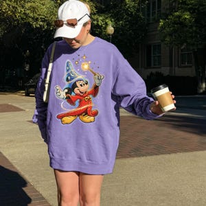 May include: A purple sweatshirt featuring a graphic of Mickey Mouse as a wizard, holding a wand with sparkling stars. The sweatshirt is long-sleeved and oversized. The image also shows a white baseball cap and sunglasses.