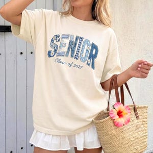 May include: A cream-colored short-sleeve t-shirt featuring the word SENIOR in large, blue, patchwork-style letters with floral and gingham patterns, and the text Class of 2027 printed in a smaller blue script font underneath the main graphic.