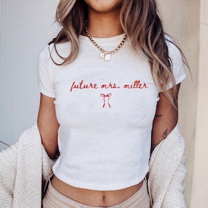 May include: White t-shirt with the red text "future mrs. miller" and a red bow graphic. The shirt has a crew neck and short sleeves. A gold chain necklace is visible.