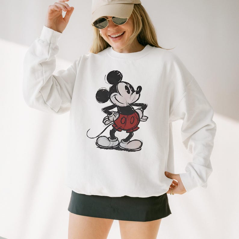 May include: White sweatshirt featuring a distressed graphic of Mickey Mouse in his classic pose. The cartoon character is depicted in black, red, and white. The sweatshirt has long sleeves and a crew neck.