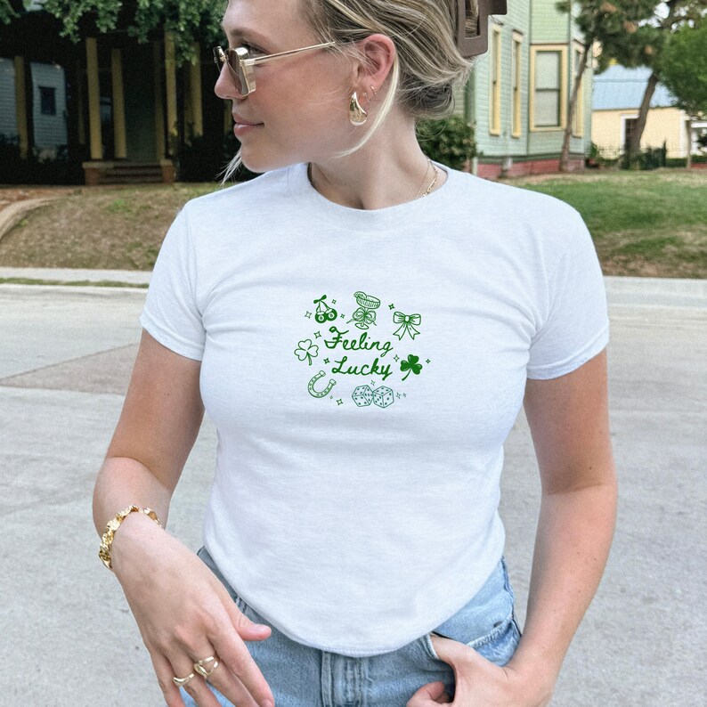 May include: White t-shirt with a crew neck and short sleeves. The shirt features a green graphic design with the words "Feeling Lucky" and lucky charms such as cherries, a horseshoe, a bow, a four-leaf clover, and dice.