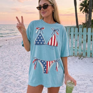 May include: A light blue short-sleeve t-shirt featuring a graphic print of a bikini with an American flag pattern. The bikini top has stars on one side and stripes on the other, while the bikini bottom features a split star and stripe design.