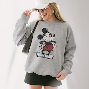May include: A gray sweatshirt featuring a distressed graphic of Mickey Mouse in his classic pose. The cartoon character is depicted in black, red, and white. The sweatshirt is a crew neck style.