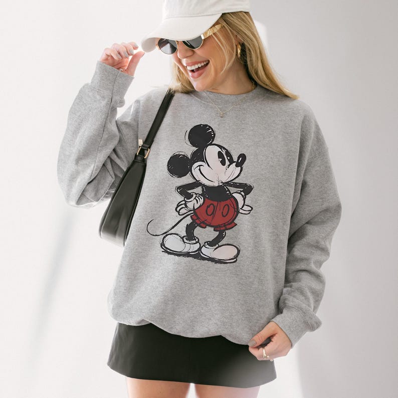 May include: A gray sweatshirt featuring a distressed graphic of Mickey Mouse in his classic pose. The cartoon character is depicted in black, red, and white. The sweatshirt is a crew neck style.