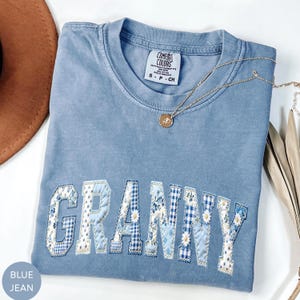 May include: A blue jean colored t-shirt with the word "GRANNY" appliqued across the front. The letters are made of various blue and white floral and patterned fabrics. A gold necklace with a pendant is draped across the shirt.