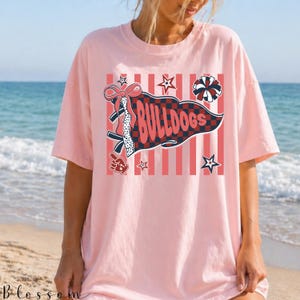May include: A light pink t-shirt featuring a graphic print of a red and navy blue checkered pennant with the text BULLDOGS. The design includes a red bow, a pom-pom, a foam finger, and stars over a background of vertical red and white stripes.