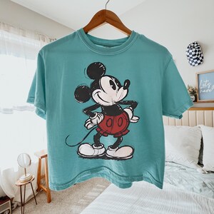 May include: A teal-colored t-shirt featuring a graphic of Mickey Mouse. The cartoon character is depicted in black, white, and red, with a smiling expression. The shirt is hanging on a wooden hanger.