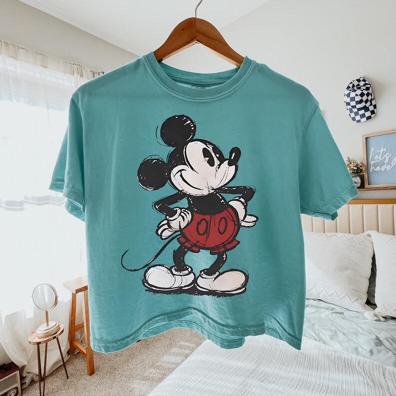 May include: A teal-colored t-shirt featuring a graphic of Mickey Mouse. The cartoon character is depicted in black, white, and red, with a smiling expression. The shirt is hanging on a wooden hanger.