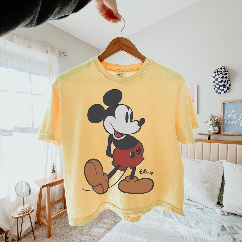 May include: Yellow t-shirt featuring a large graphic of Mickey Mouse. The cartoon character is depicted in black, white, red, and brown. The Disney logo is printed in black on the lower right side of the shirt.