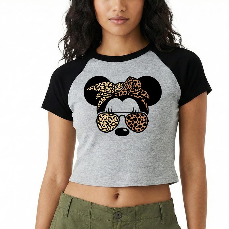 Op de afbeelding: Een grijze raglan crop top met zwarte mouwen met een Minnie Mouse afbeelding. De afbeelding bevat zwarte muizenoren, een luipaardprint strik, zonnebril en een neus. Het shirt is een casual, stijlvol kledingstuk.