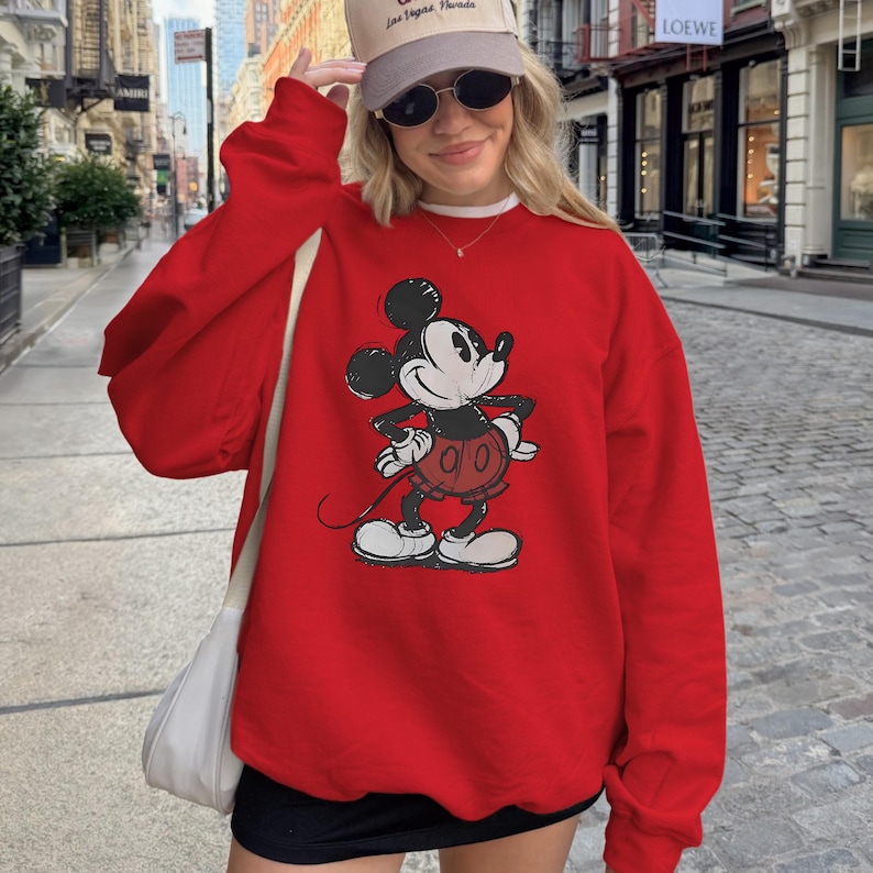 May include: A red sweatshirt featuring a classic Mickey Mouse graphic. The cartoon character is depicted in black, white, and red. The sweatshirt is a solid red color and has long sleeves. The design is a vintage style.