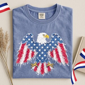 May include: A blue Comfort Colors t-shirt featuring an embroidered American flag eagle design. The eagle has a white head, yellow beak and talons, and wings with a blue star field and red and white stripes. The shirt is folded.