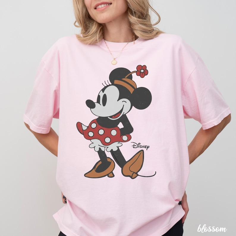 Może przedstawiać: Jasnor&oacute;żowa koszulka z grafiką Myszki Minnie. Myszka nosi czerwoną sp&oacute;dnicę w kropki, brązowe buty i kapelusz z kwiatkiem. Logo Disneya jest nadrukowane na koszulce.
