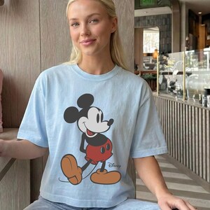 May include: Light blue t-shirt featuring a graphic of Mickey Mouse in a walking pose. The cartoon character is depicted in black, red, and tan colors. The Disney logo is printed on the lower right side of the shirt.