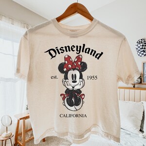 K&ouml;nnte beinhalten: Cremefarbenes T-Shirt mit einem Minnie-Mouse-Motiv mit roter Schleife und Kleid. Das Shirt tr&auml;gt den Text "Disneyland" &uuml;ber dem Motiv, mit "est. 1955 CALIFORNIA" darunter. Das Shirt h&auml;ngt an einem Holzb&uuml;gel.