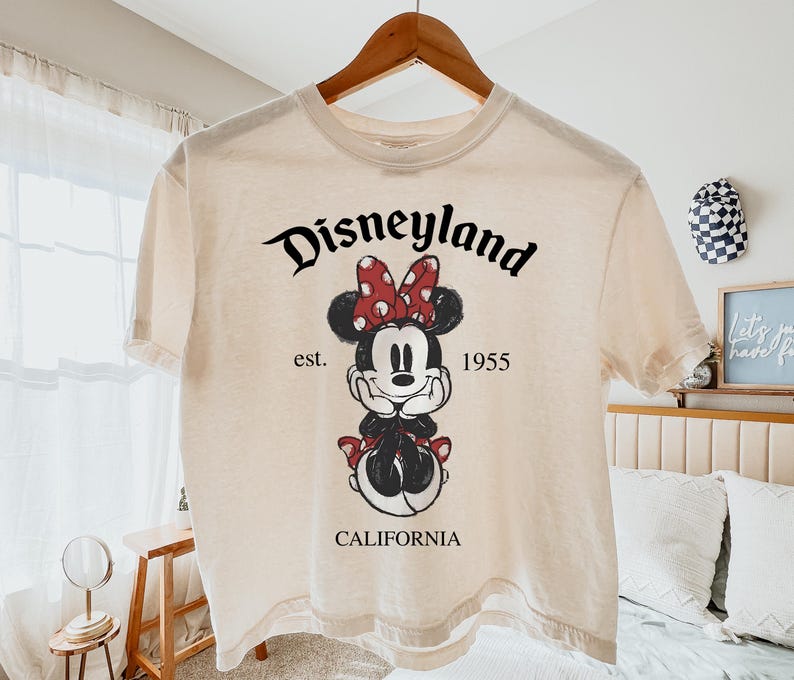 K&ouml;nnte beinhalten: Cremefarbenes T-Shirt mit einem Minnie-Mouse-Motiv mit roter Schleife und Kleid. Das Shirt tr&auml;gt den Text "Disneyland" &uuml;ber dem Motiv, mit "est. 1955 CALIFORNIA" darunter. Das Shirt h&auml;ngt an einem Holzb&uuml;gel.