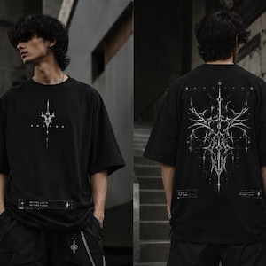 May include: A black short-sleeved t-shirt featuring a small white graphic on the chest and a large, intricate, symmetrical white abstract design on the back. The shirt includes small rectangular text boxes with the words BEYOND LIMITS BEYOND FLESH.