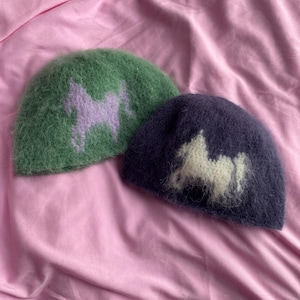 Puede incluir: Dos gorros difusos. Uno es verde con un diseño de caballo lila, y el otro es azul marino con un diseño de caballo blanco. Los gorros se muestran sobre un fondo de tela rosa.