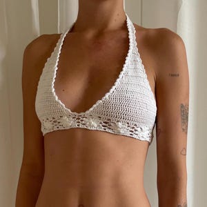 Può includere: Top all'uncinetto bianco fatto a mano. Il top presenta un delicato motivo floreale lungo il bordo inferiore e un laccetto all'americana. Il lavoro all'uncinetto è intricato, con un aspetto strutturato. Il top è progettato per essere indossato come capo d'abbigliamento.