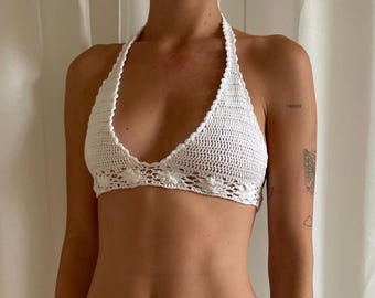 May include: A white, handmade crochet halter top. The top features a delicate floral pattern along the bottom edge and a halter neck tie. The crochet work is intricate, with a textured appearance. The top is designed to be worn as a garment.