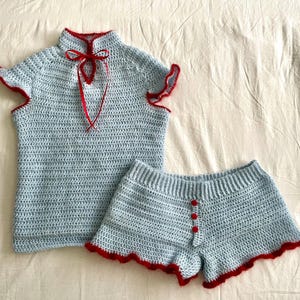 May include: A light blue crochet pajama set. The top has a high collar with a red trim and a red ribbon tie. The shorts have red trim and three red buttons. The set is handmade.