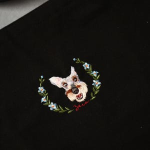 May include: Black fabric with an embroidered Schnauzer dog design. The dog is surrounded by a wreath of blue flowers and green leaves. The dog has a white face, brown nose, and open mouth.