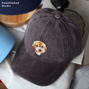 Pode incluir: Boné de beisebol cinza escuro com uma imagem bordada de um cão pequeno, marrom e branco. O boné tem uma aba curva e um botão na parte superior. O texto "Pawstitched Studio" é visível no canto superior esquerdo.