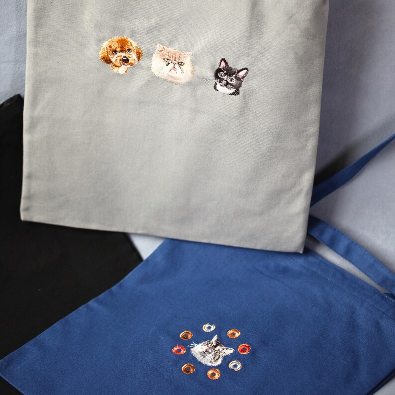 May include: Gray tote bag embroidered with a poodle, a Persian cat, and a tuxedo cat. A blue tote bag with a cat embroidered in the center, surrounded by orange and brown circles. A black tote bag is also visible.