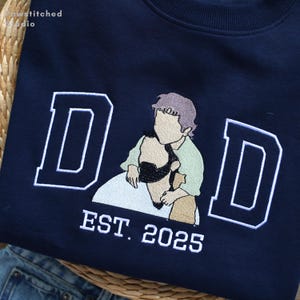 Pode incluir: Camisola azul marinho com a palavra "DAD" em letras brancas e uma ilustração bordada de uma pessoa segurando uma criança. O texto "EST. 2025" está abaixo da palavra "DAD". A camisola é feita de um material macio.