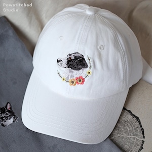 Può includere: Cappellino da baseball bianco con un ritratto di cane ricamato. Il cappellino presenta un cane bianco e nero all'interno di una ghirlanda floreale. Ha una visiera curva e un bottone sulla parte superiore. Il marchio "Pawstitched Studio" è visibile.