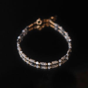 Op de afbeelding: Een delicate armband met twee strengen kleine, gespikkelde witte en zwarte kralen, afgewisseld met goudkleurige afstandhouders. De armband is vastgemaakt met een gouden sluiting tegen een donkere achtergrond.