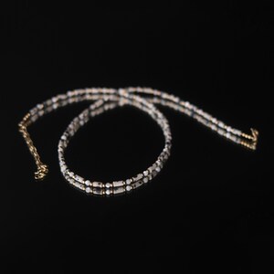 Op de afbeelding: Een delicate ketting met kleine, gespikkelde kralen in zwart, wit en goud. De ketting heeft een dubbele streng met een goudkleurige ketting en sluiting. De kralen zijn gelijkmatig verdeeld, wat zorgt voor een verfijnde en elegante uitstraling.