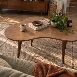 May include: A kidney-shaped wooden coffee table with three tapered legs. On the surface sits a white ceramic mug on a coaster, a stack of books topped with a small brown bowl containing keys, a potted green plant, and a dark glass candle.