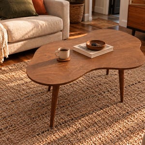 May include: A kidney-shaped wooden coffee table with three tapered legs. The tabletop features a smooth, medium-brown finish and holds a white ceramic mug on a coaster, a rectangular book, and a small round wooden bowl containing keys.