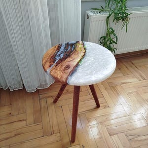 Pode incluir: Uma mesa auxiliar redonda com tampo de resina epóxi branca e marrom e três pernas de madeira. A mesa está sobre um piso de madeira.