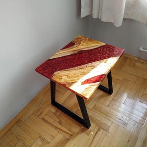 Könnte beinhalten: Kleiner, quadratischer Beistelltisch mit einzigartigem Design. Die Tischplatte kombiniert helles Holz und tiefrotes Harz, getragen von schwarzen Metallbeinen. Die Tischabmessungen betragen ca. 50 cm Länge, 50 cm Breite und 46 cm Höhe.