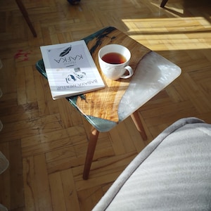Puede incluir: Pequeña mesa auxiliar con tapa de madera y detalles de resina. Un libro titulado "Kafka" y una taza de té están sobre la mesa. La mesa tiene tres patas de madera y está sobre un suelo de madera. Medidas: aprox. 60 cm.