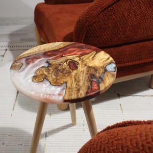 Puede incluir: Una pequeña mesa auxiliar redonda con un diseño único. La tapa de la mesa presenta una combinación de madera, resina blanca y roja. La mesa tiene tres patas de madera. La mesa está junto a un sillón color óxido.