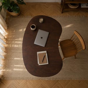 May include: A dark brown kidney-shaped wooden desk with a silver laptop, a white ceramic mug, a small glass candle holder, and a spiral-bound notebook with a black pen placed on top. A wooden spindle-back chair is positioned next to the desk.