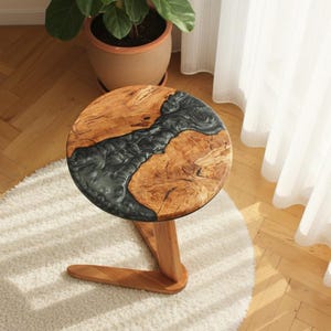 Puede incluir: Mesa auxiliar redonda de madera con una tapa de vetas de madera natural y un diseño de río de resina epoxi gris oscuro. La mesa tiene una sola pata de madera curvada. La mesa tiene aproximadamente 46 cm de diámetro.