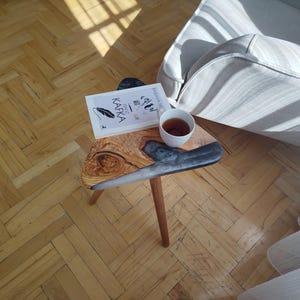 May include: A small, triangular wooden side table with a live edge and a dark grey resin inlay. A book and a cup of tea sit on the table. The table has three wooden legs and is placed on a wooden floor.