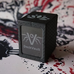 Könnte beinhalten: Eine schwarze, rechteckige DeckVault-Aufbewahrungsbox mit strukturierter Oberfläche und einem Lotusblüten-Symbol auf der Oberseite. Die Vorderseite zeigt ein stilisiertes V-Logo mit einer Augengrafik und dem hellgrauen Schriftzug DeckVault.