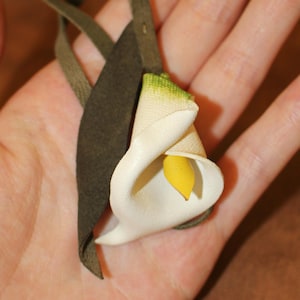 Può includere: Un ciondolo a forma di calla in pelle fatto a mano con un petalo bianco e stami gialli. Il fiore è attaccato a un cinturino in pelle verde scuro. Il ciondolo è tenuto nella mano di una persona.