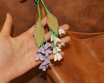 May include: A close-up of a floral accessory with a leather leaf and flower design. The flowers are in shades of white and lavender, with green stems. The background is a brown leather bag.