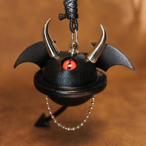 Puede incluir: Un llavero de cuero negro con un ojo rojo, cuernos plateados y alas. El llavero está unido a una cadena plateada con un dije de flecha plateada.